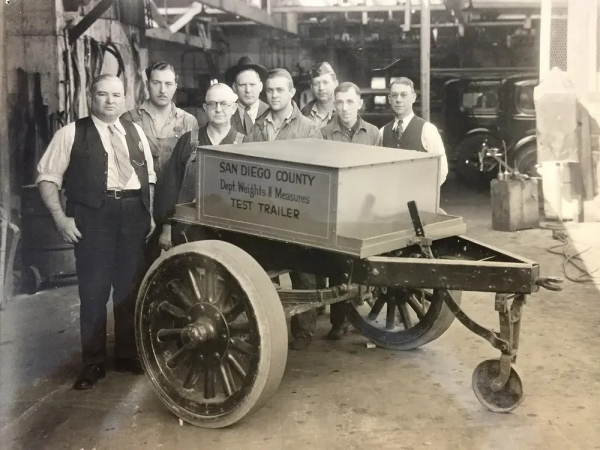 Black and white photo from the 1930s or 40s of San Diego County Weights & Measures inspectors posing beside an equipment trailer marked "San Diego County Dept. Weights & Measures Test trailer."