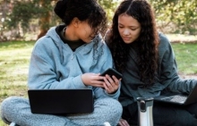 Two young women sitting on grass, reading from a phone.