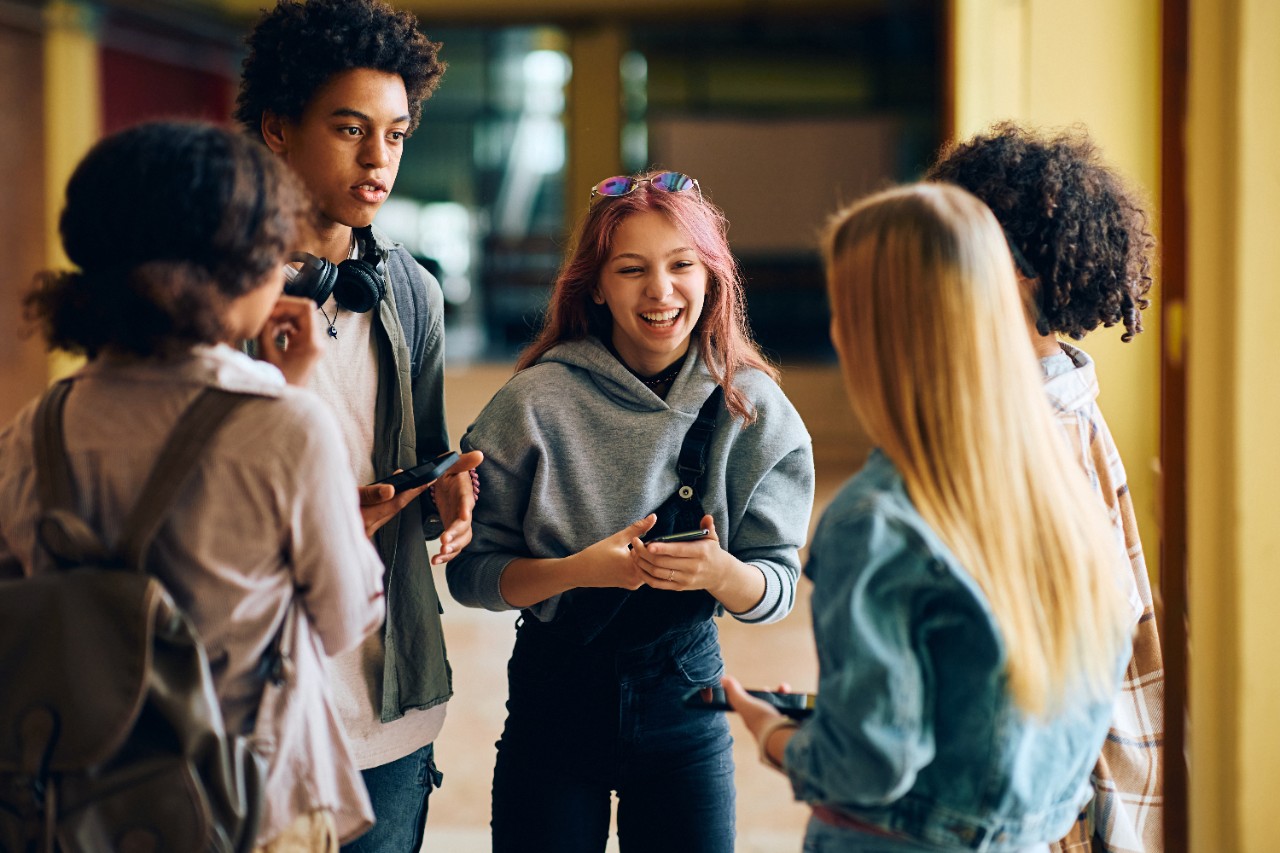 Happy teenage girl laughing while using cell phone and communicating with her friends in hallway at high school.