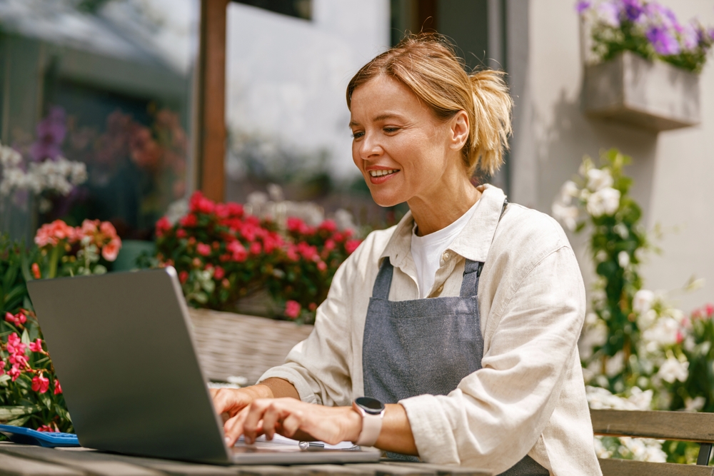 Small flower shop owner florist wearing apron working on laptop and taking online orders in store 