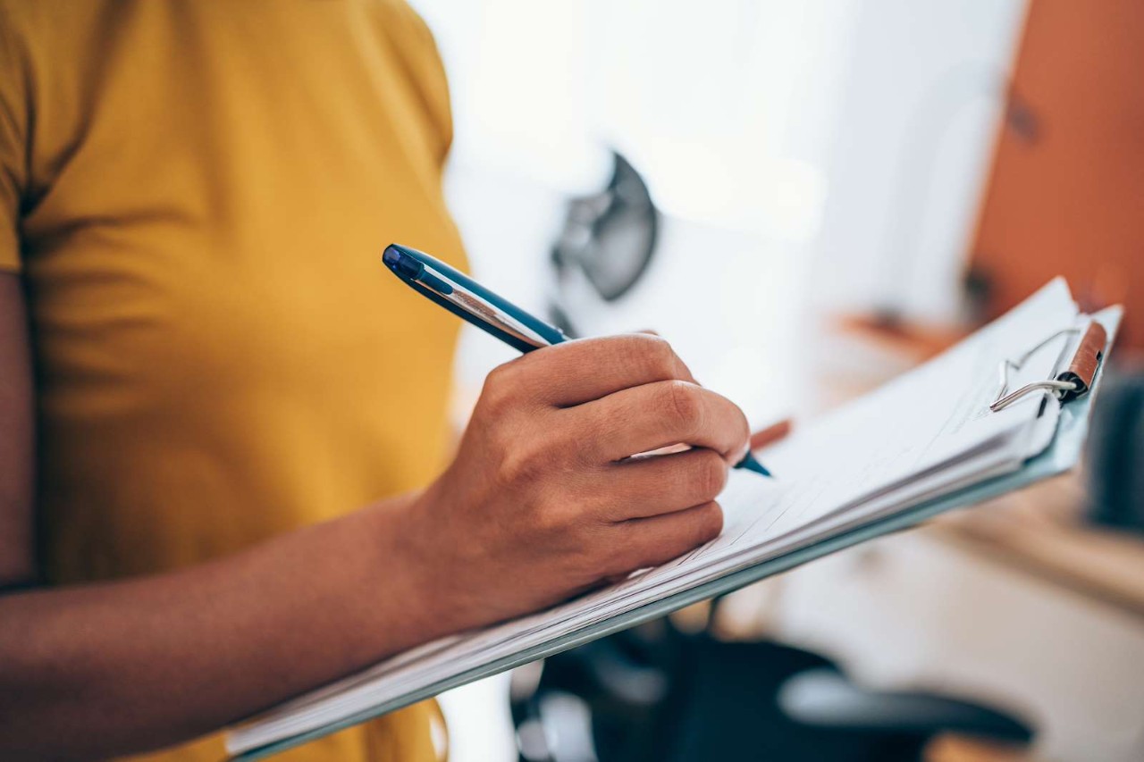 A person holding a pen and a clipboard with papers attached