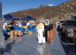HHW event - people dressed in hazmat gear, cars lined up between cones, hazardous waste in barrels
