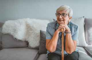 Older adult woman sitting on a couch while smiling and resting their chin on their hands and cane 