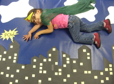 Photo of CCS child, dressed up as a superhero and laying on a floor poster to create the illusion of flying over buildings..