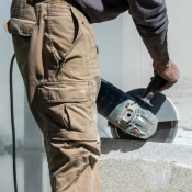 Person wearing dusty work clothes uses a handheld power saw to cut through a concrete or stone surface outdoors. 