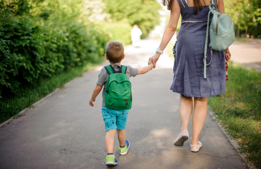 An adult holding hands with a child as they walk on a path carrying backpacks.