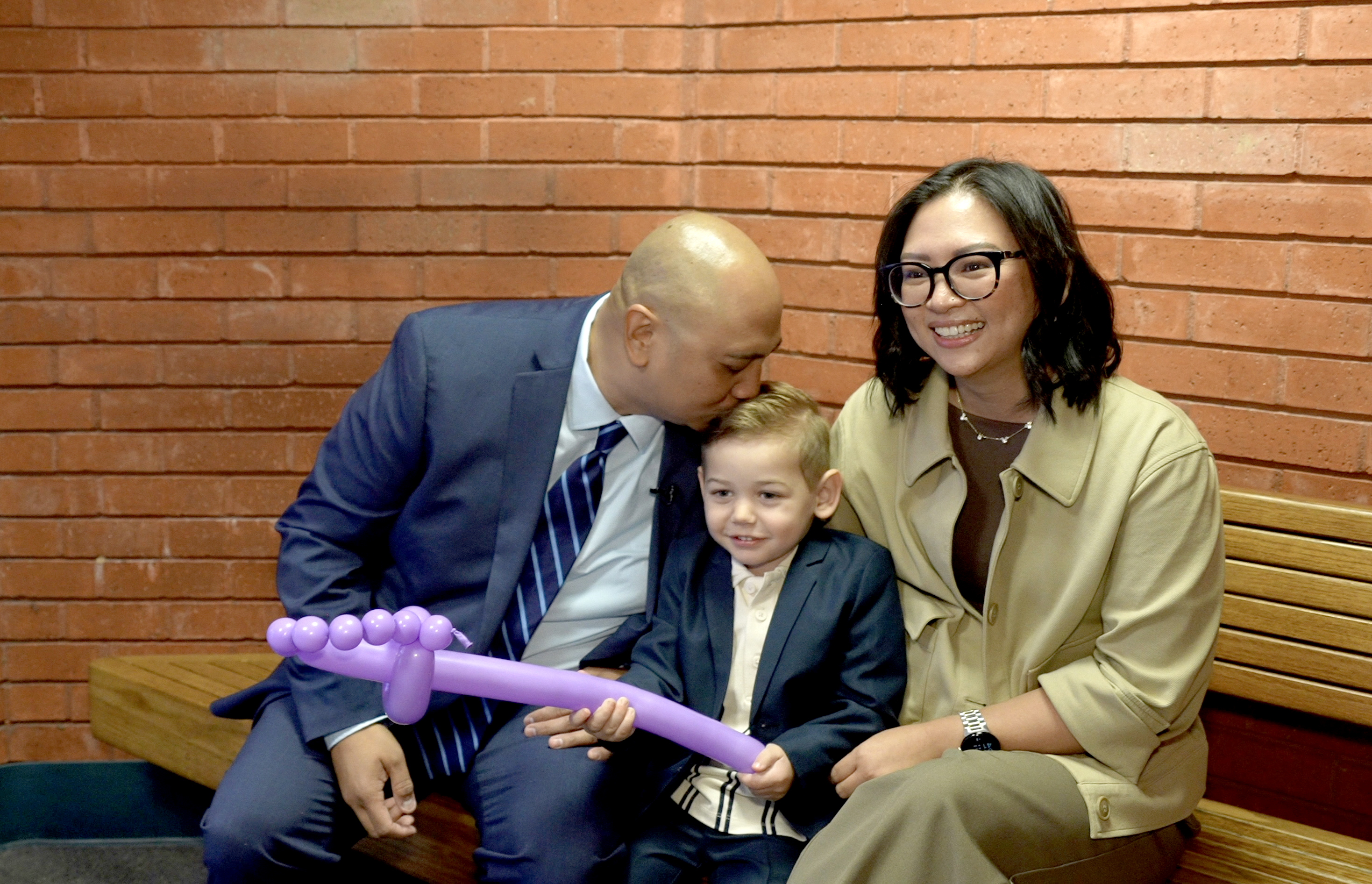 Family of three sitting on a bench. Father on the left, wearing a suit, leaning over kissing the top of his blond, adopted son's head. The son is holding a light purple balloon shaped as a sword. The mother, sitting and smiling on the right, is wearing a tan suit and eyeglasses.